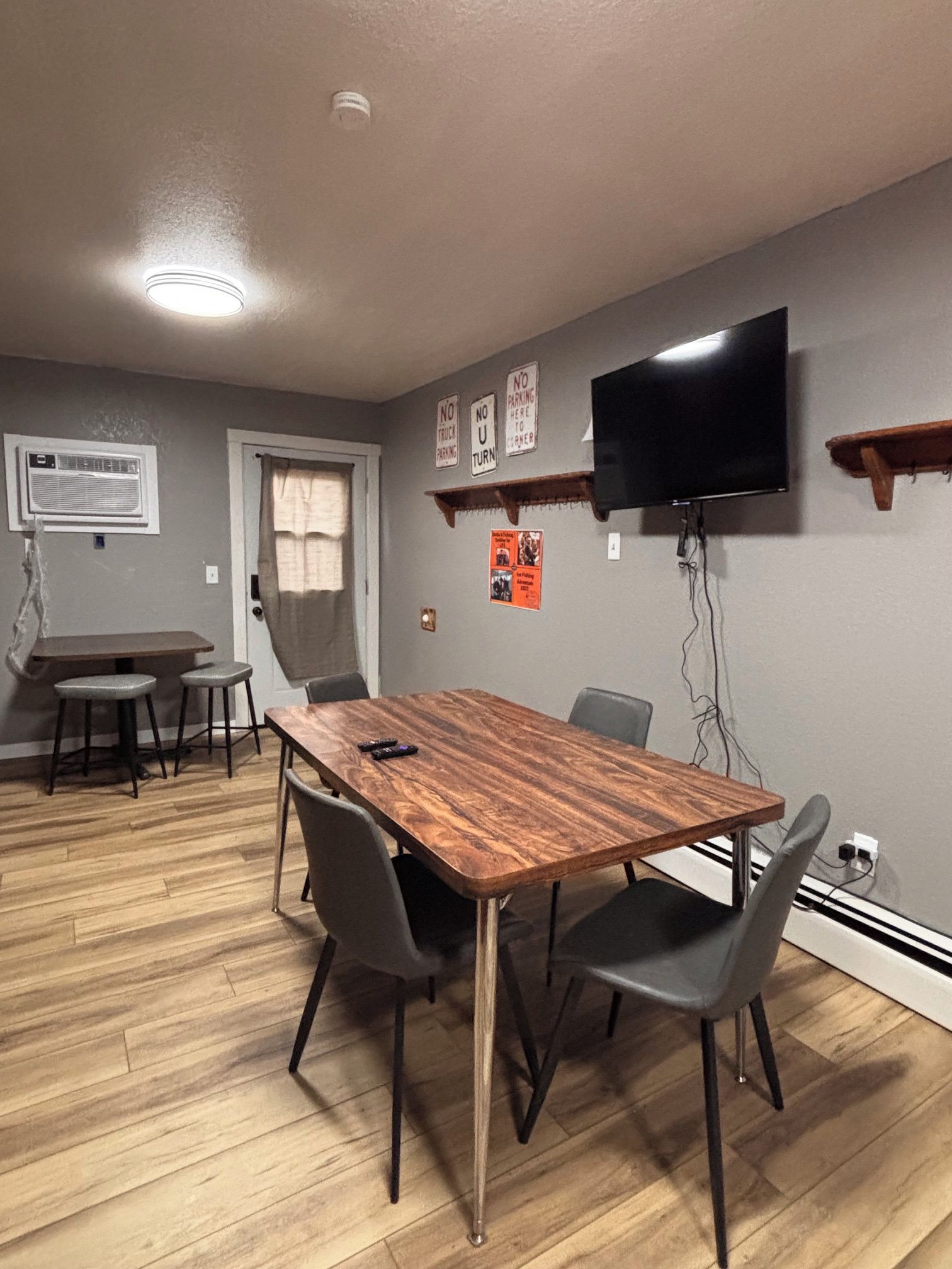 A dining area with a wooden table, four chairs, a television mounted on the wall, and a window with curtains.