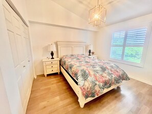 A bedroom with a bed, nightstands, a hanging light fixture, a window with blinds, and a door. - Naples Coastal Condo with Pool – 2 Bed / 2 Bath – 20 Minutes from the Beach. (Naples)