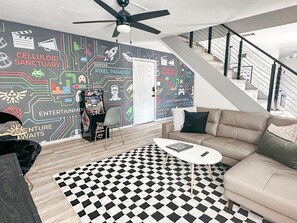 A modern living room with a checkered floor, a sofa, a coffee table, and a wall with gaming and entertainment-themed graphics. - Gamer's Day Den - Walk to Kyle Field (College Station)