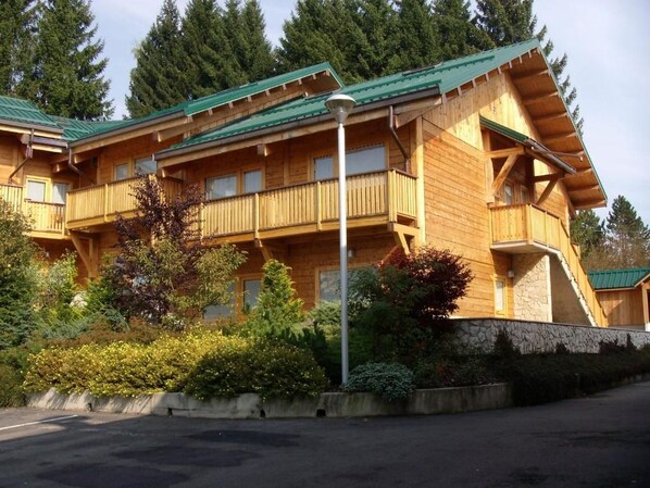 A multi-story wooden building with balconies, surrounded by trees and shrubs.