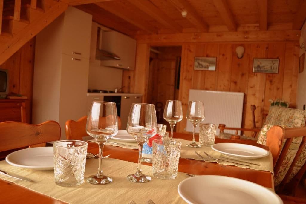 A dining table set with wine glasses and plates, wooden walls, and a kitchen in the background.
