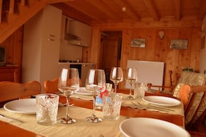 A dining table set with wine glasses and plates, wooden walls, and a kitchen in the background.
