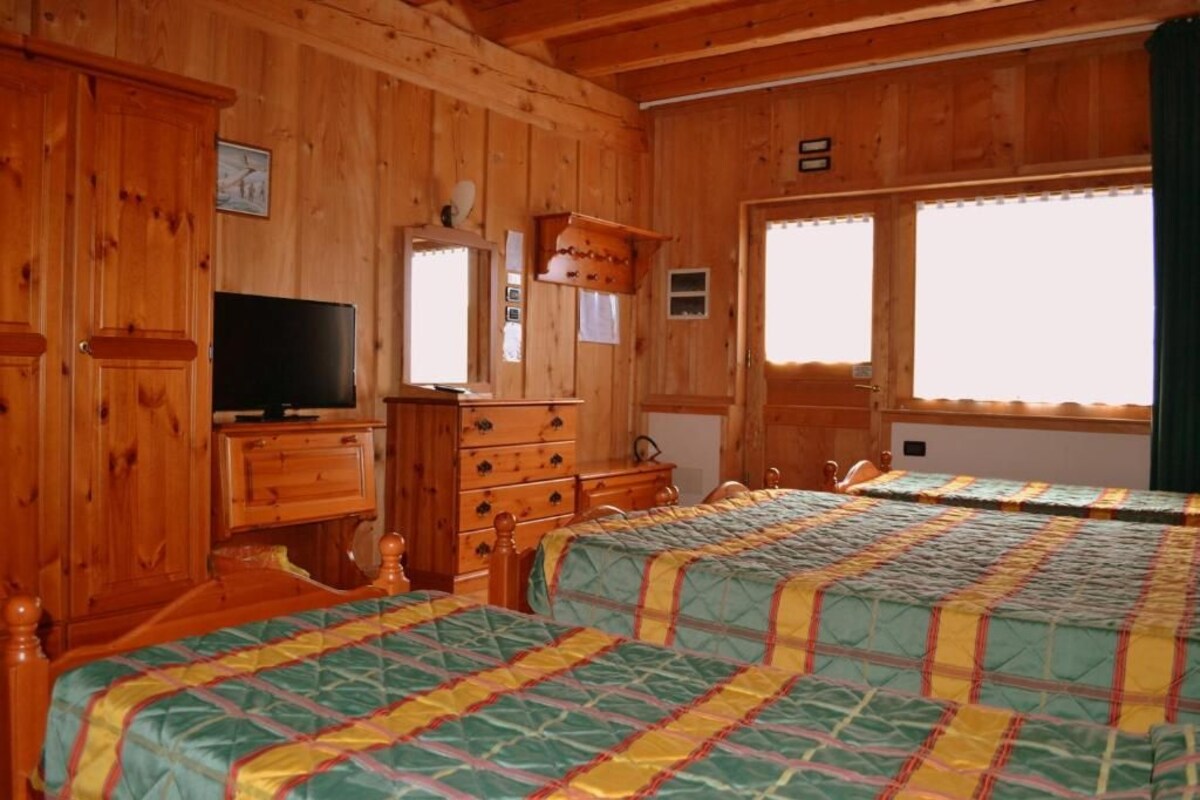 A wooden cabin-style room with two beds, a dresser, a television, and large windows.