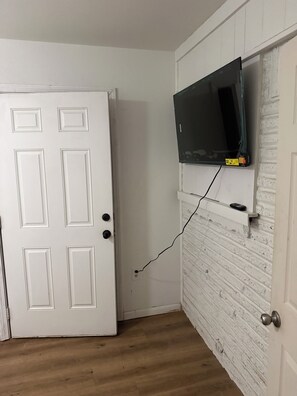 A room with a white door, a mounted television on a brick-patterned wall, and a wooden floor. - Beautiful 4-bedroom house in enchanting West Orange with AC (West Orange)