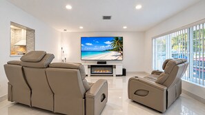 A modern living room with a flat-screen TV, leather reclining chairs, a fireplace, and large windows offering a view of the outdoors. - Private Pool | BBQ | Outdoor Dining (Hollywood)