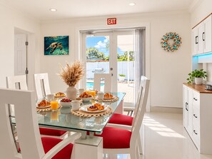 A modern dining area with a glass table, red chairs, breakfast items, and a view of a pool and greenery outside. - Private Pool | BBQ | Outdoor Dining (Hollywood)