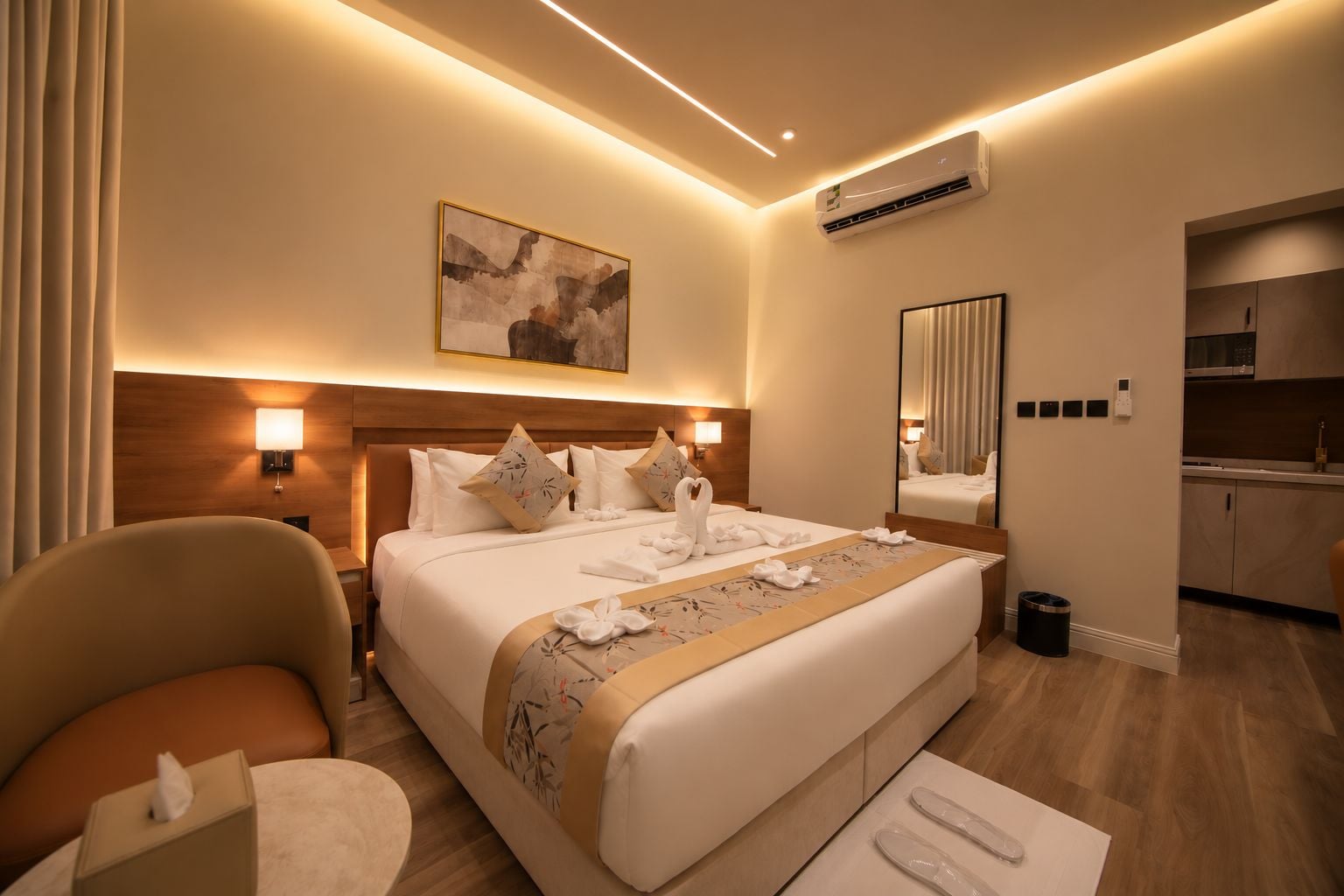 A hotel room with a large bed, bedside lamps, a painting, an air conditioning unit, a mirror, a small kitchenette, and a trash bin.