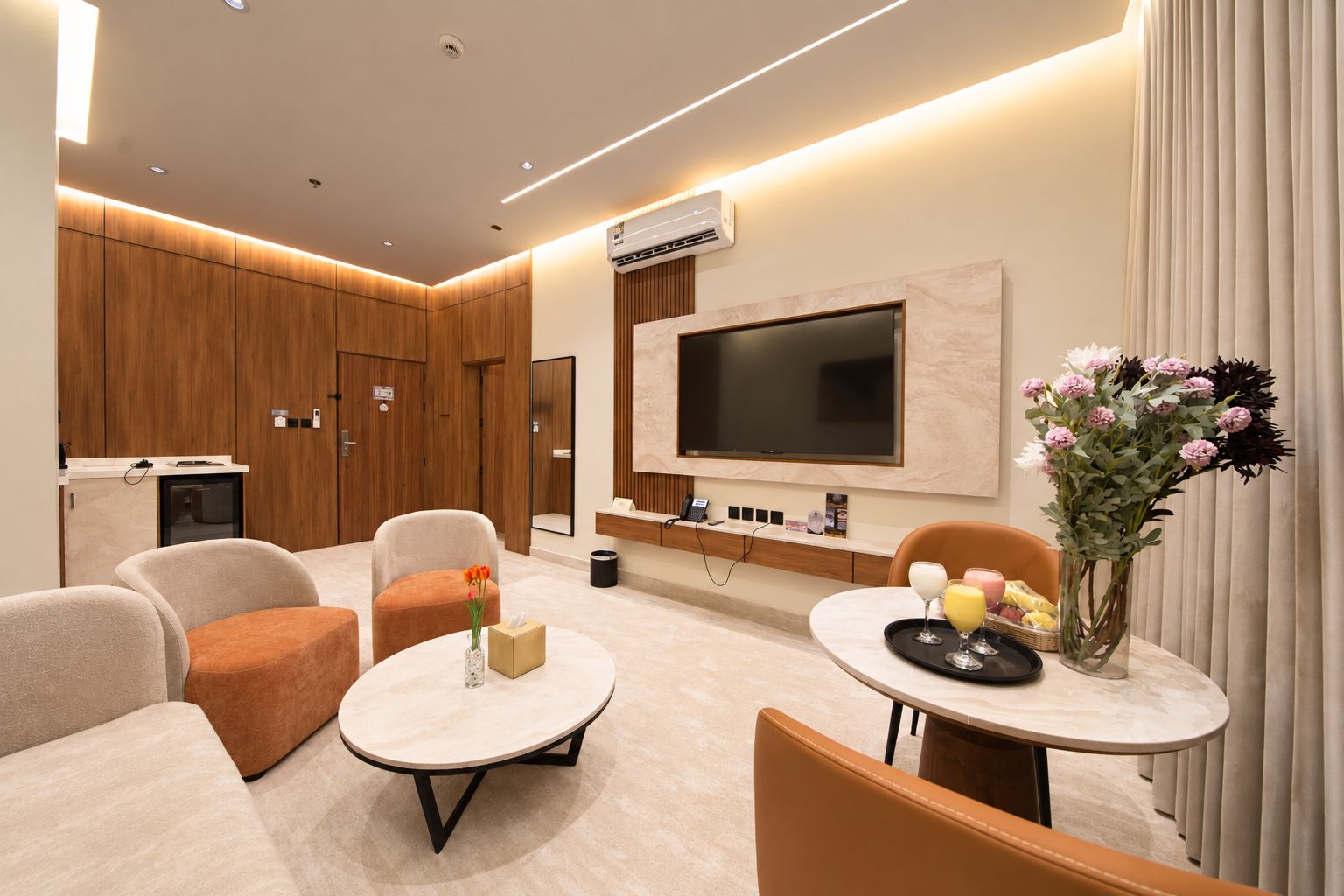 A modern hotel room with a flat-screen TV, comfortable seating, a small table with a vase of flowers, and a minibar.