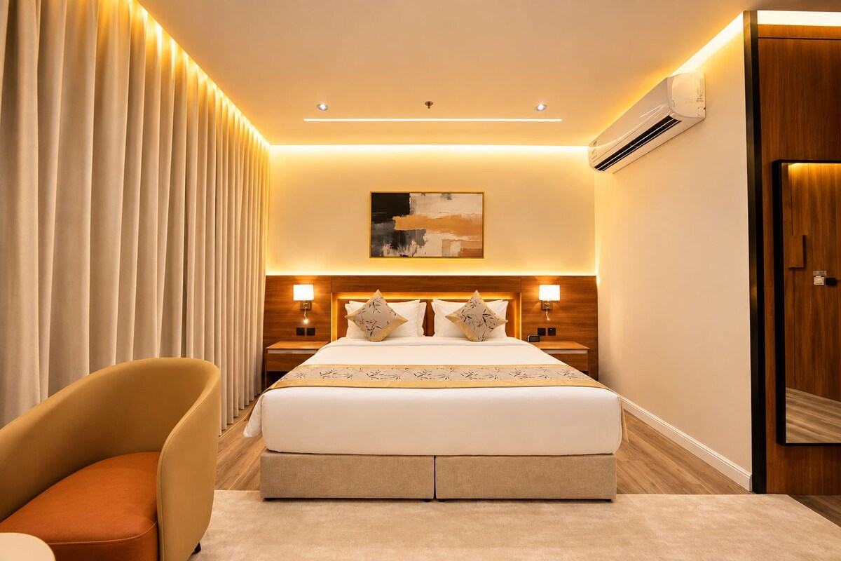 A hotel room with a large bed, bedside lamps, a headboard with a painting, an air conditioning unit, and a wooden door.