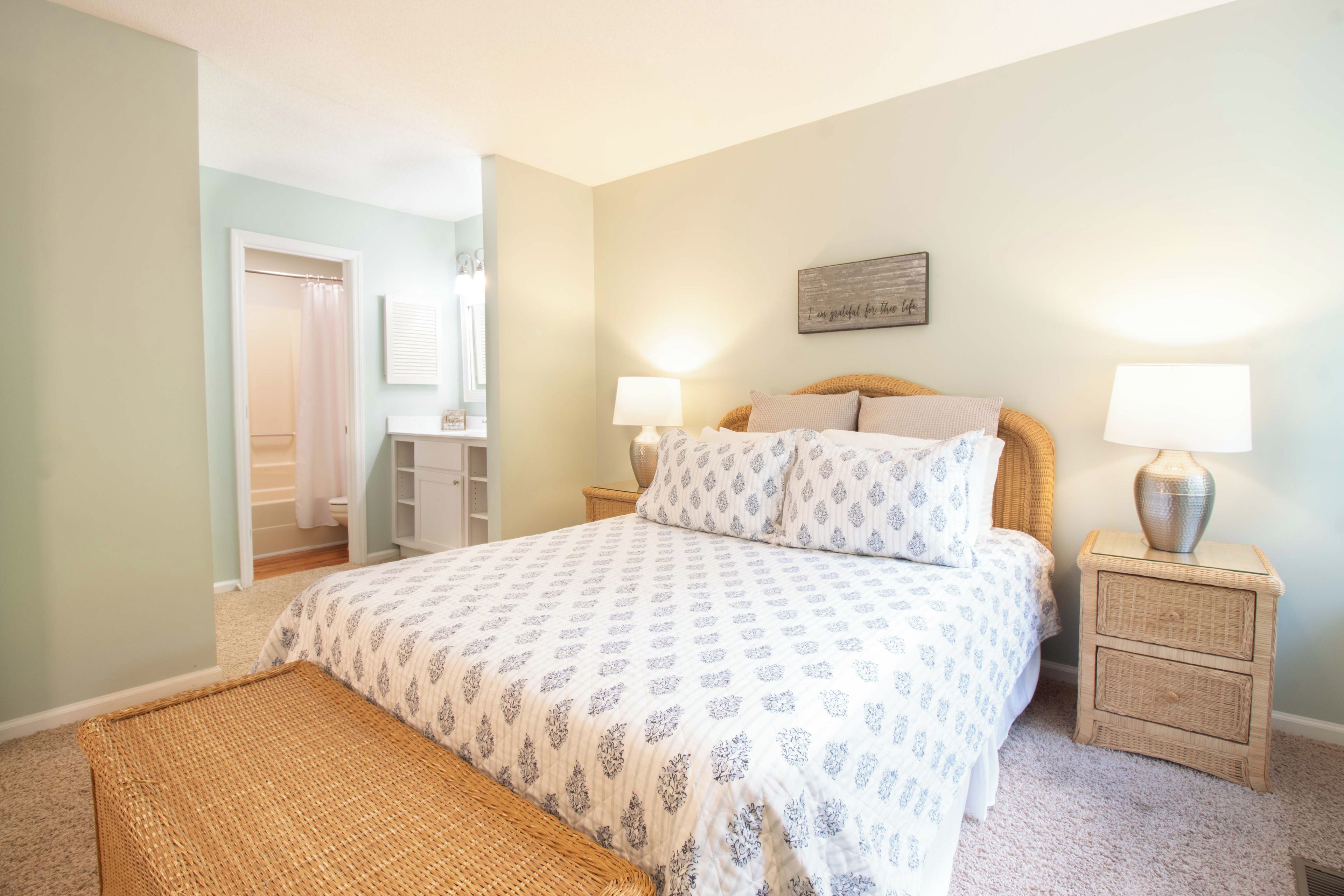 A bedroom with a bed, two bedside tables, a nightstand, a wicker bench, a wall-mounted lamp, and a door leading to a bathroom.