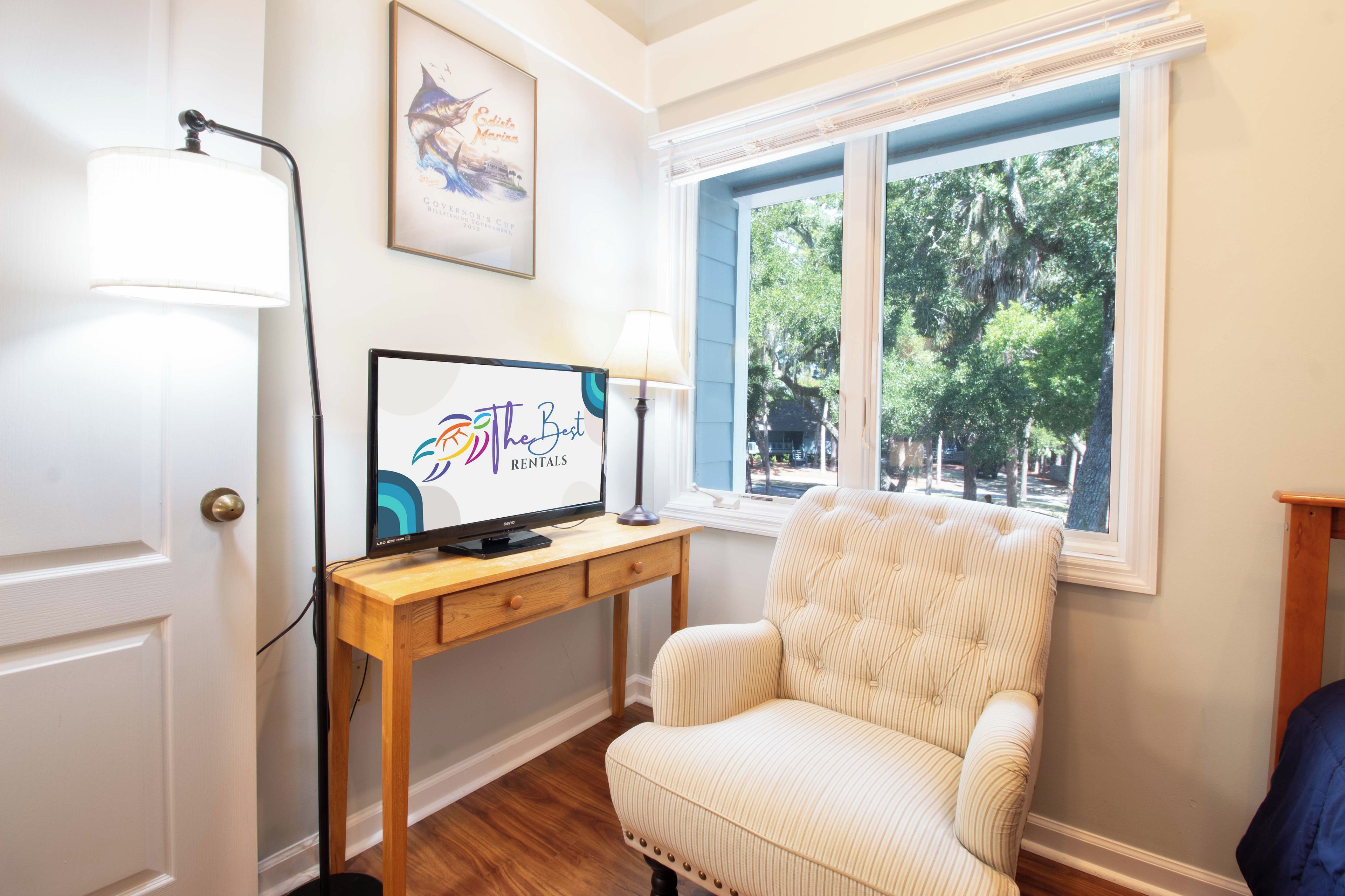 A room with a television on a wooden desk, a striped armchair, and a window with a view of trees.
