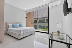 A modern bedroom with a bed, a glass table with a vase, and a view of a building through a glass door. - Distrito Corazón: 2BR, Balconies & Rooftop Pool (Playa del Carmen)
