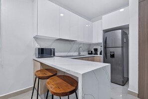 A modern kitchen with white cabinetry, a marble countertop, a built-in microwave, a stainless steel refrigerator, and two wooden stools. - Distrito Corazón: 1BR, 2 Baths & Rooftop Pool (Playa del Carmen)