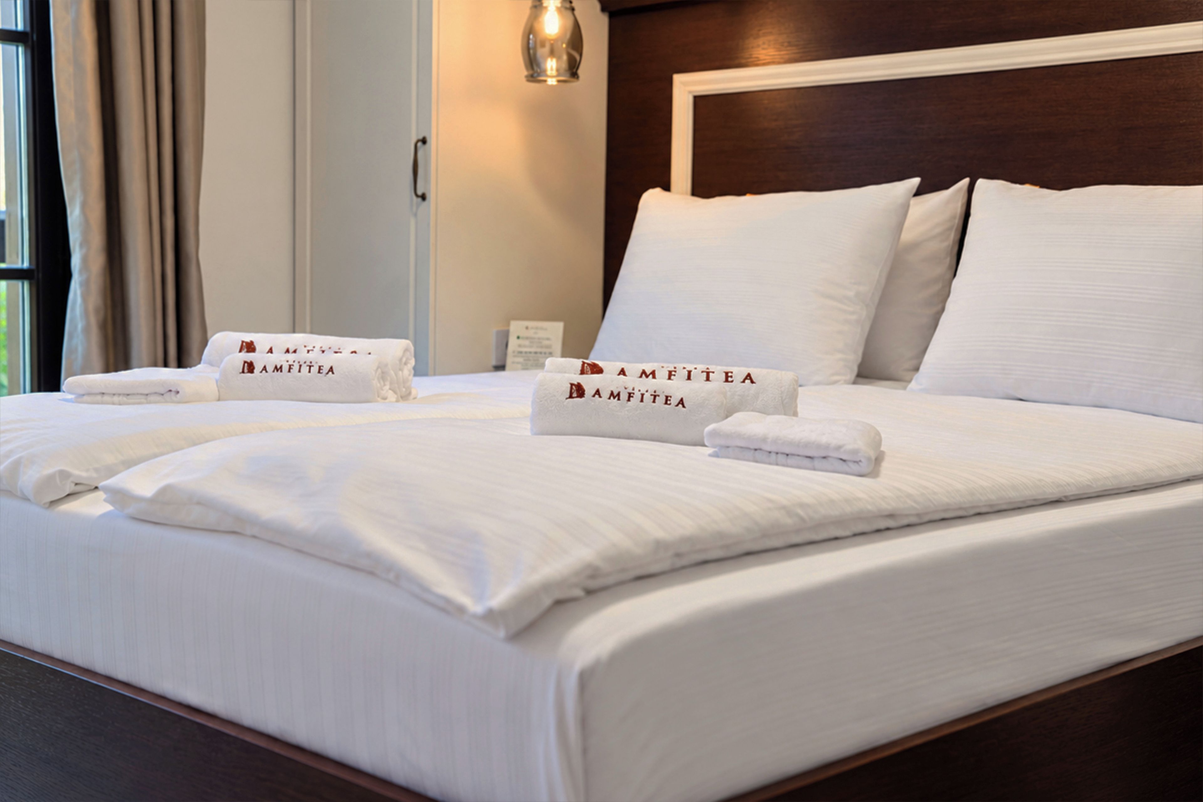 A neatly made bed with white linens and towels branded 'AMFITEA' in a hotel room.