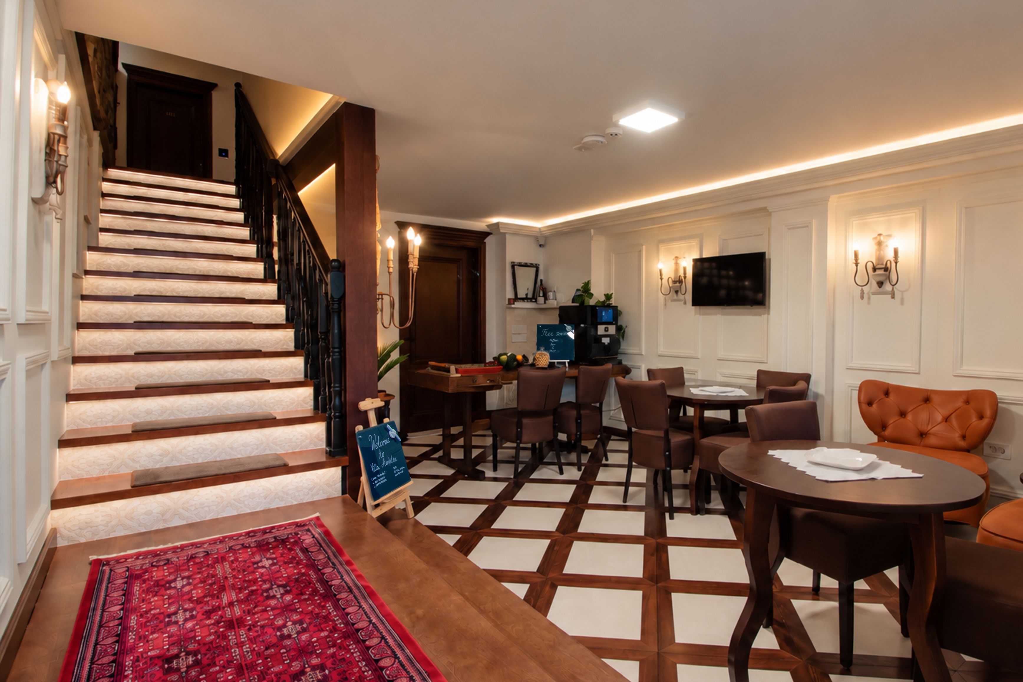 A hotel lobby with a staircase, a reception desk, a dining area with tables and chairs, and a television mounted on the wall.