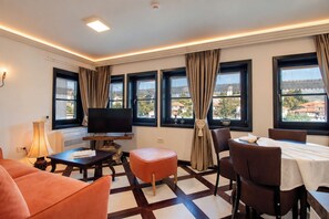 Une chambre d’hôtel équipée d’une télévision, d’une table à manger, de chaises, d’un canapé et de grandes fenêtres avec des rideaux.