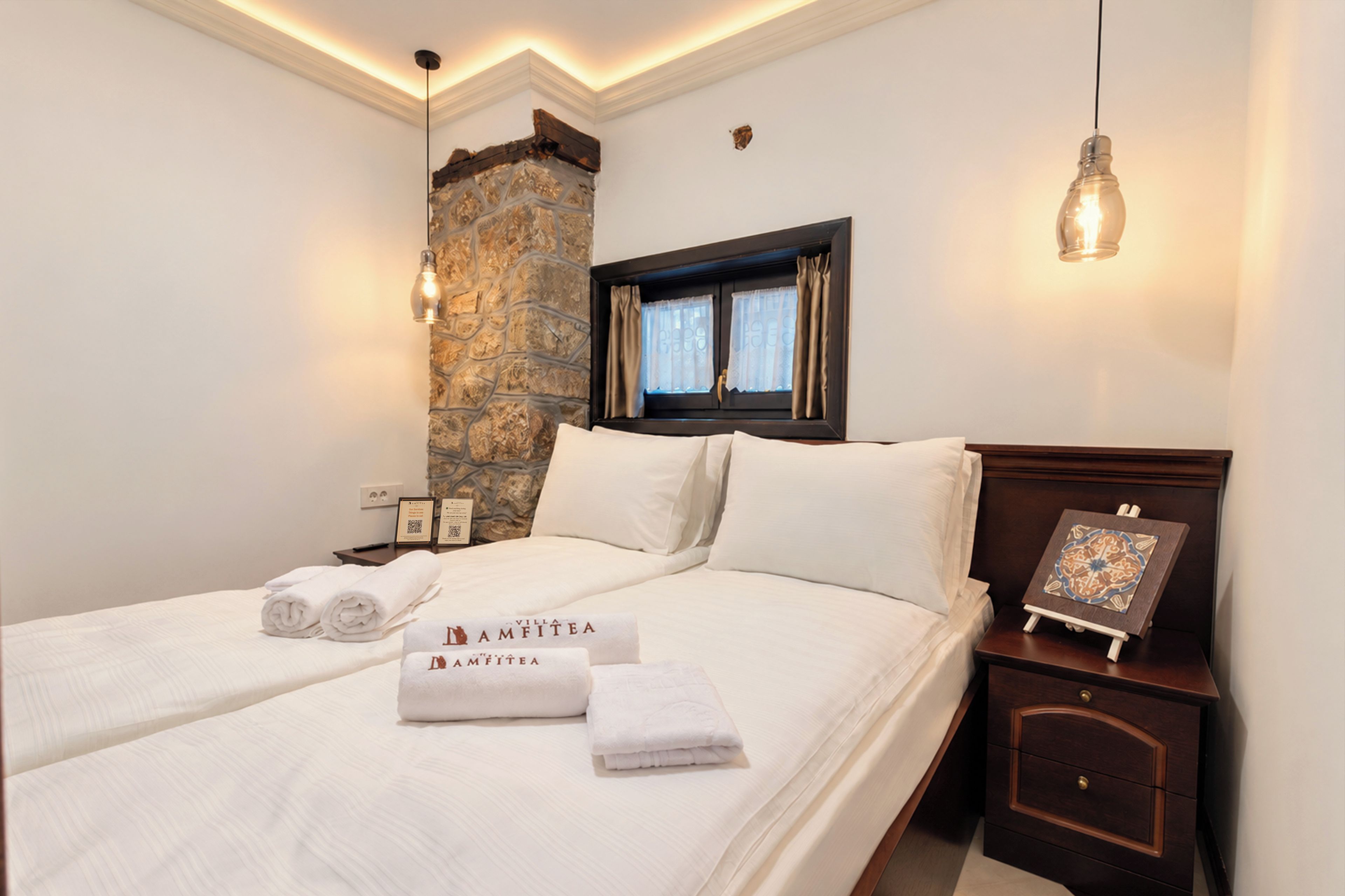Habitación de hotel con dos camas, un muro decorativo de piedra, una mesita de noche con un cuadro enmarcado y una lámpara colgante.