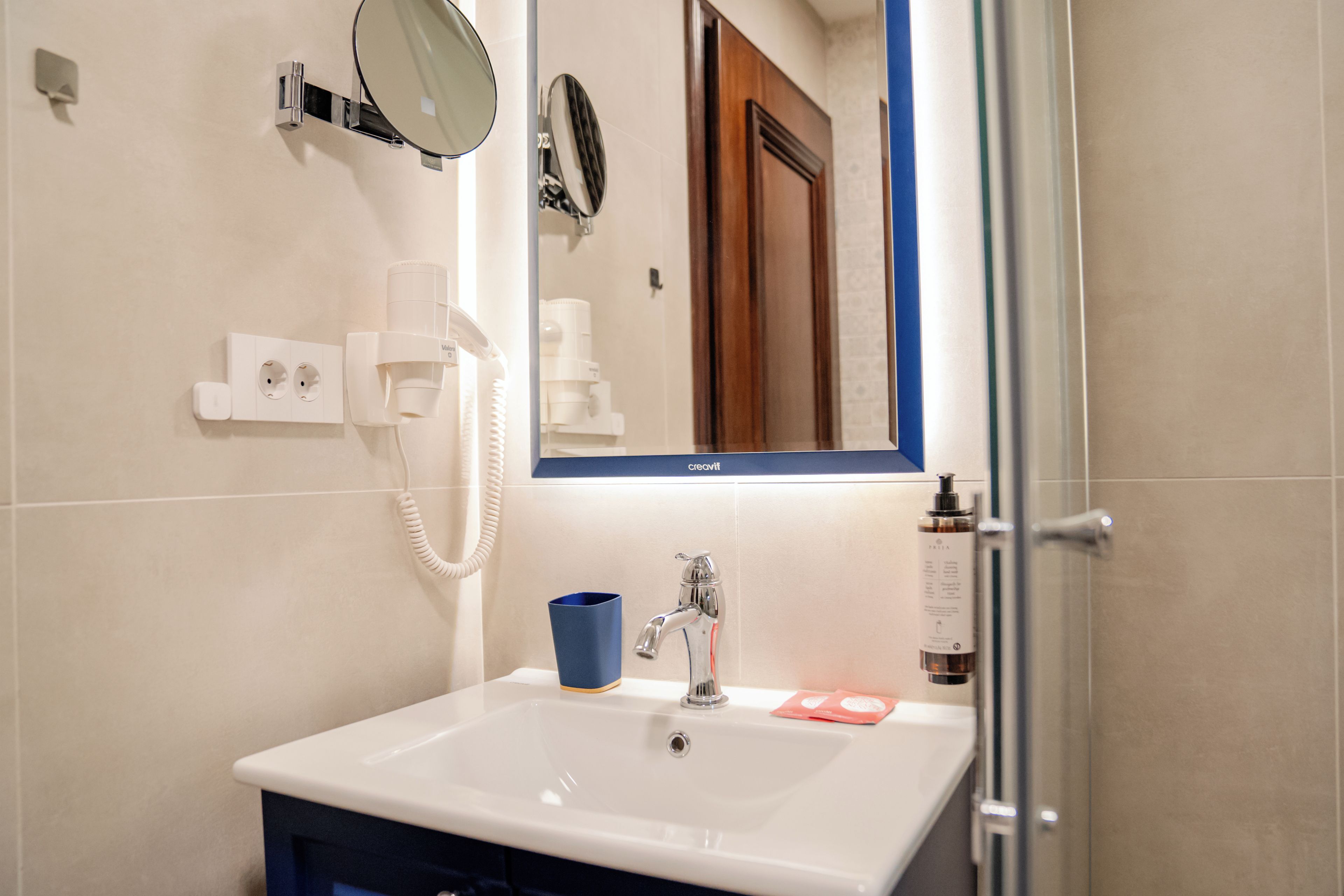 Un baño con un lavamanos blanco, un espejo, una taza azul y una puerta de ducha.