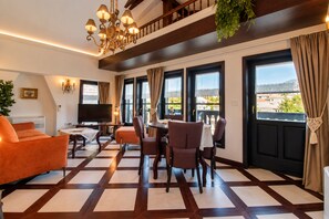 A hotel room with a checkered floor, a dining area with chairs and a table, a sofa, a TV, and large windows with curtains. - Villa Amfitea (Ohrid)