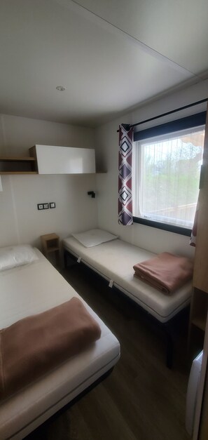 A compact room with two beds, a window with curtains, a shelf, and a small table. - Mobile home with direct beach access (Canet-en-Roussillon)