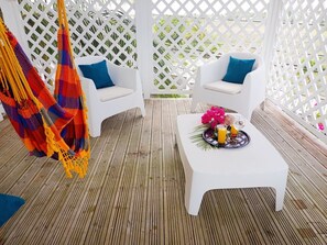 A wooden deck with two white chairs and a small white table, a colorful hammock, and a white lattice structure in the background. - Villa Azura – 4 Bedrooms, Infinity Pool, Spa & Surf | Le Helleux (Sainte-Anne)