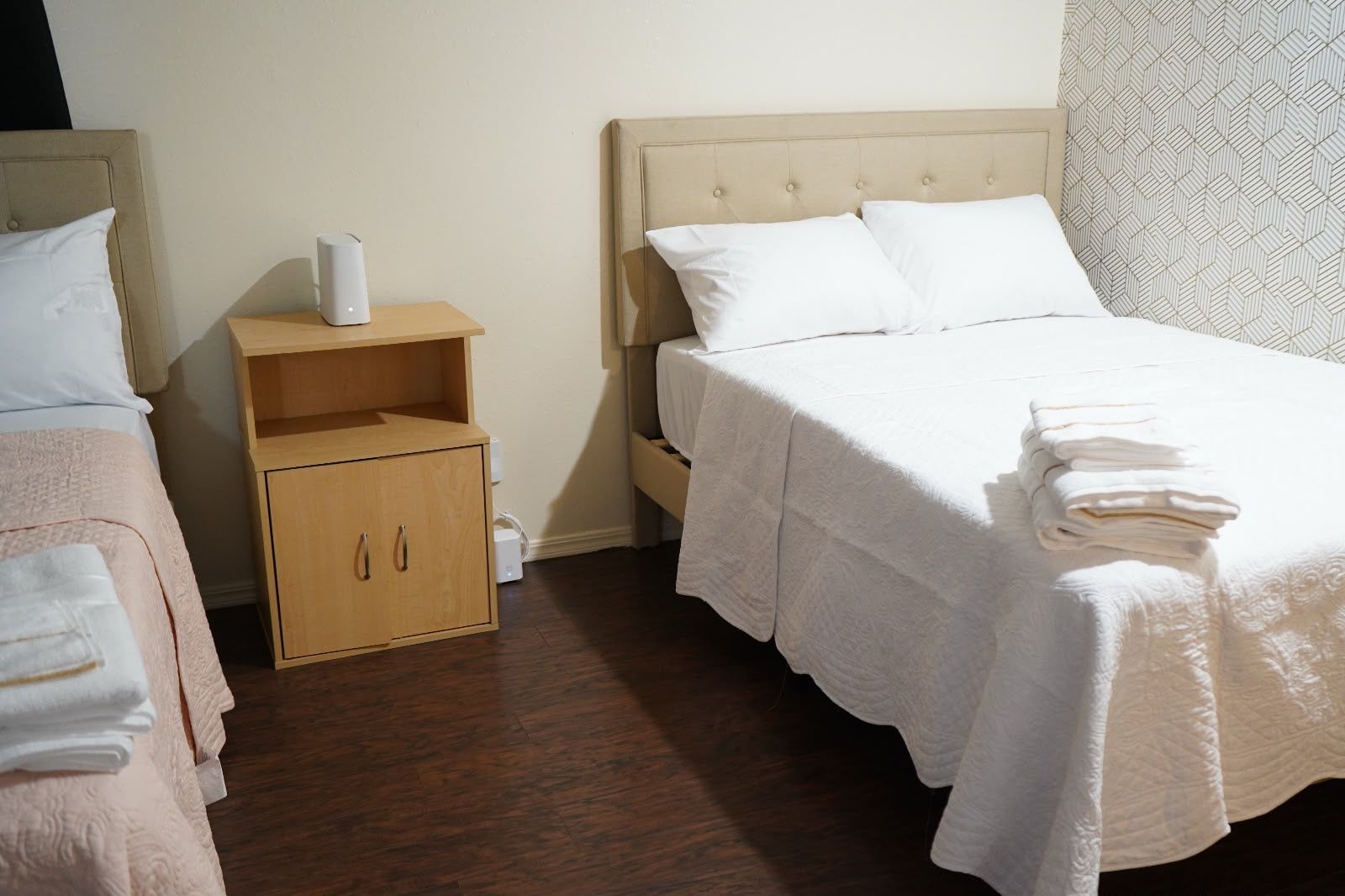 Habitación de hotel con dos camas, una mesita de noche de madera con un dispositivo blanco, un armario de madera y una pared con diseño.