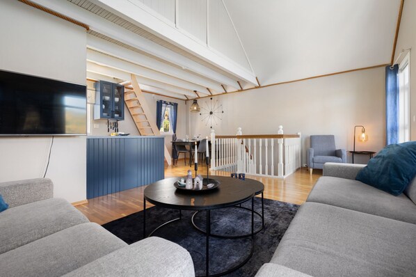 A modern living room with a large TV, a dining area, and a staircase leading to an upper level.