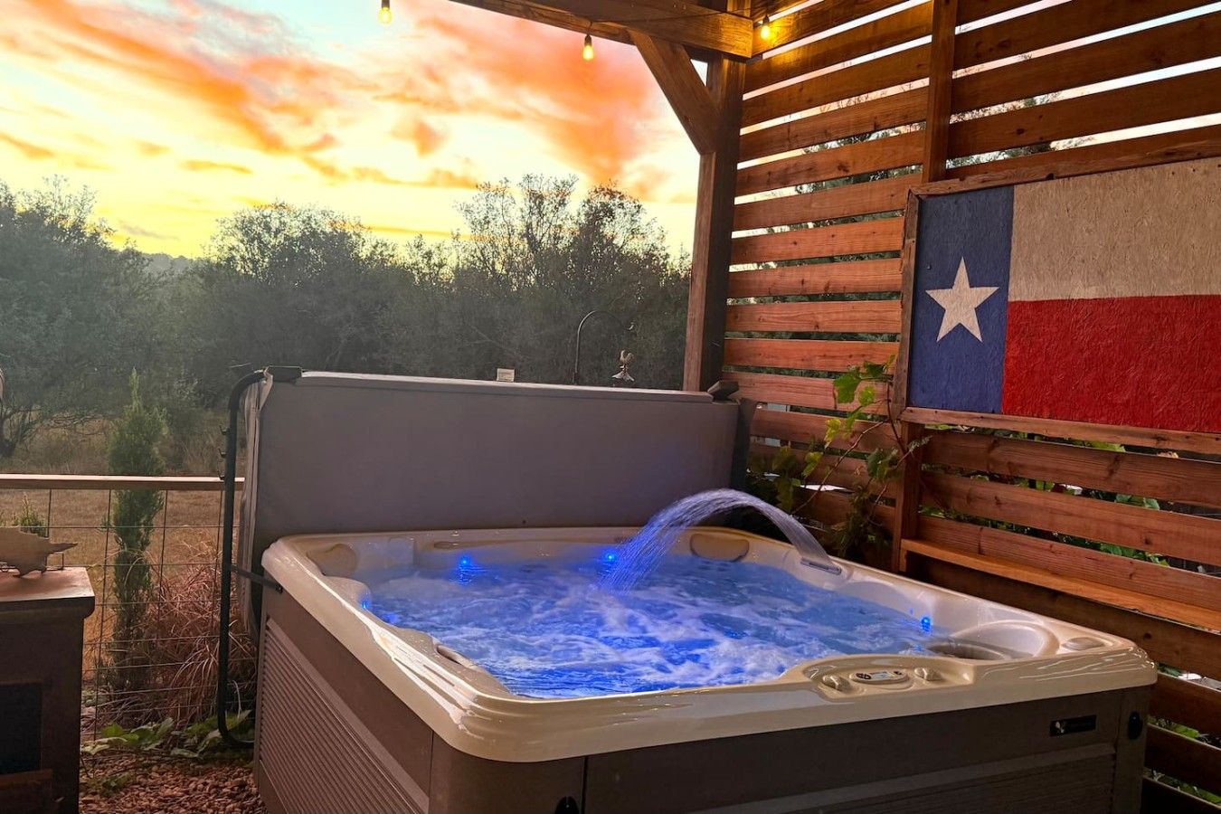 A hot tub with a Texas flag backdrop, surrounded by wooden slats and a view of trees.