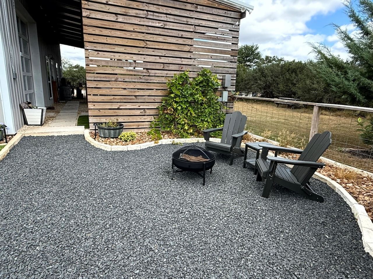 An outdoor seating area with a fire pit and Adirondack chairs, surrounded by a gravel patio and a wooden fence.