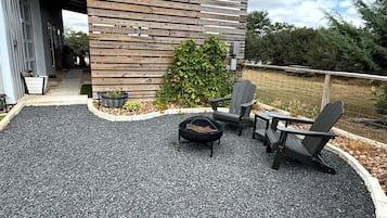 An outdoor seating area with a fire pit and Adirondack chairs, surrounded by a gravel patio and a wooden fence.