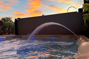 A hot tub with a water fountain feature, surrounded by a wooden deck and a sunset sky.