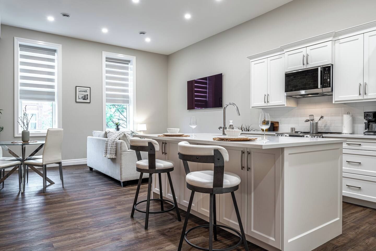 Une cuisine moderne avec des meubles blancs, un îlot central doté de tabourets, un four à micro-ondes encastré et un coin repas avec un canapé et des chaises.