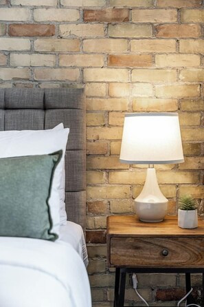 Un lit avec une tête de lit grise, un oreiller blanc et un coussin décoratif vert. Il y a une table de chevet avec une lampe et une petite plante en pot. Le mur derrière est fait de briques apparentes.