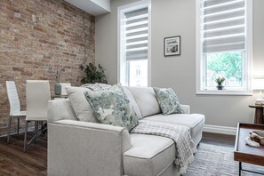 Un salon avec un mur en briques, un canapé blanc agrémenté de coussins à motifs, une table à manger avec des chaises et une fenêtre avec des stores.