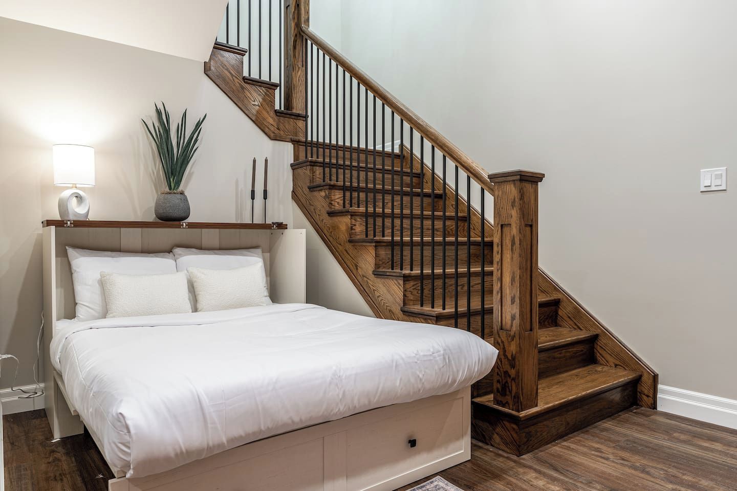 Une chambre avec un grand lit, un escalier en bois, une lampe de chevet et une plante en pot.