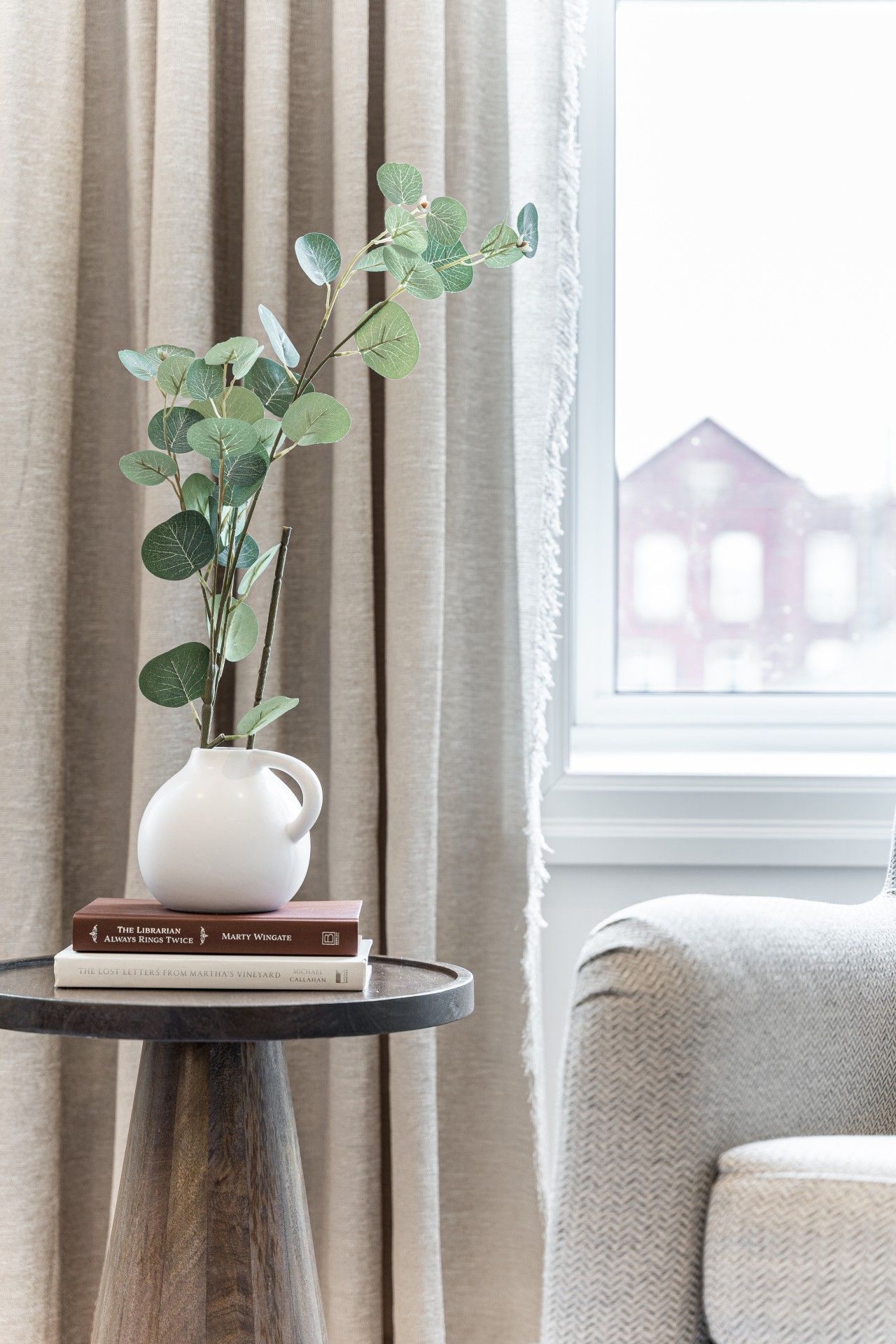 Eine weiße Vase mit grünen Blättern auf einem runden Tisch, neben einem Fenster mit Vorhängen.