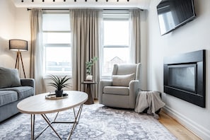 A living room with a grey sofa, a round coffee table, a grey armchair, a small side table with a plant, and a flat-screen TV mounted on the wall. - Juno F5: The Curator’s Studio (Stratford)