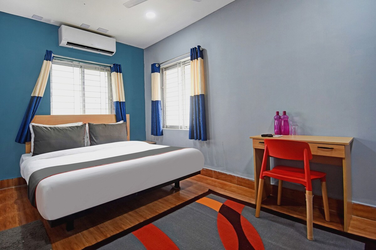 Habitación de hotel con cama, escritorio, silla y dos ventanas con cortinas.