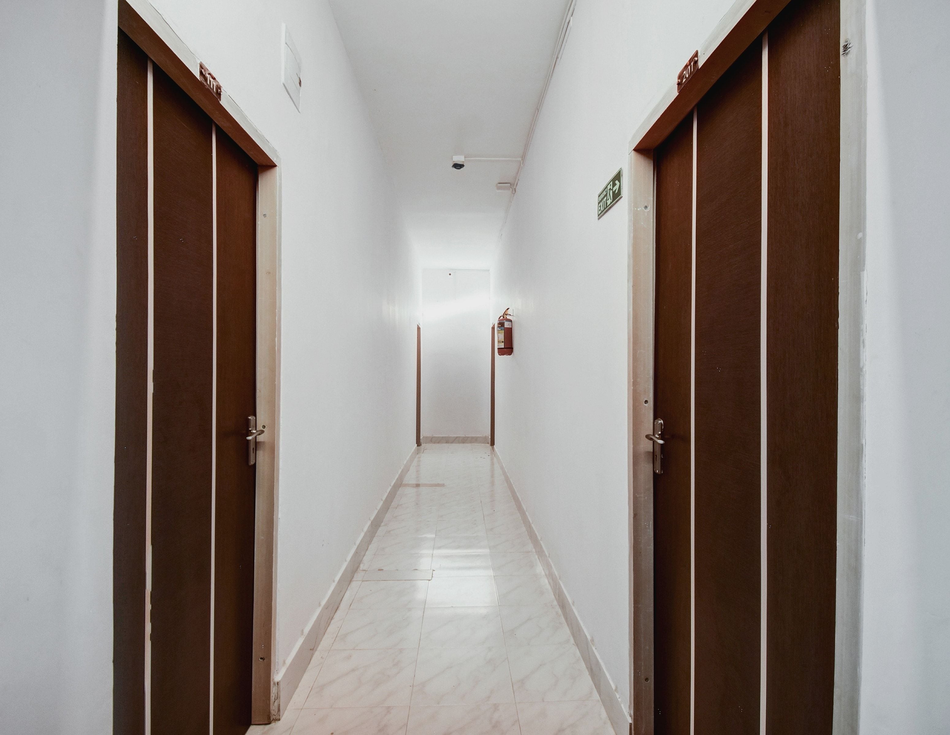 Un pasillo con puertas a ambos lados, un extintor en la pared y un letrero que indica la dirección de la salida.
