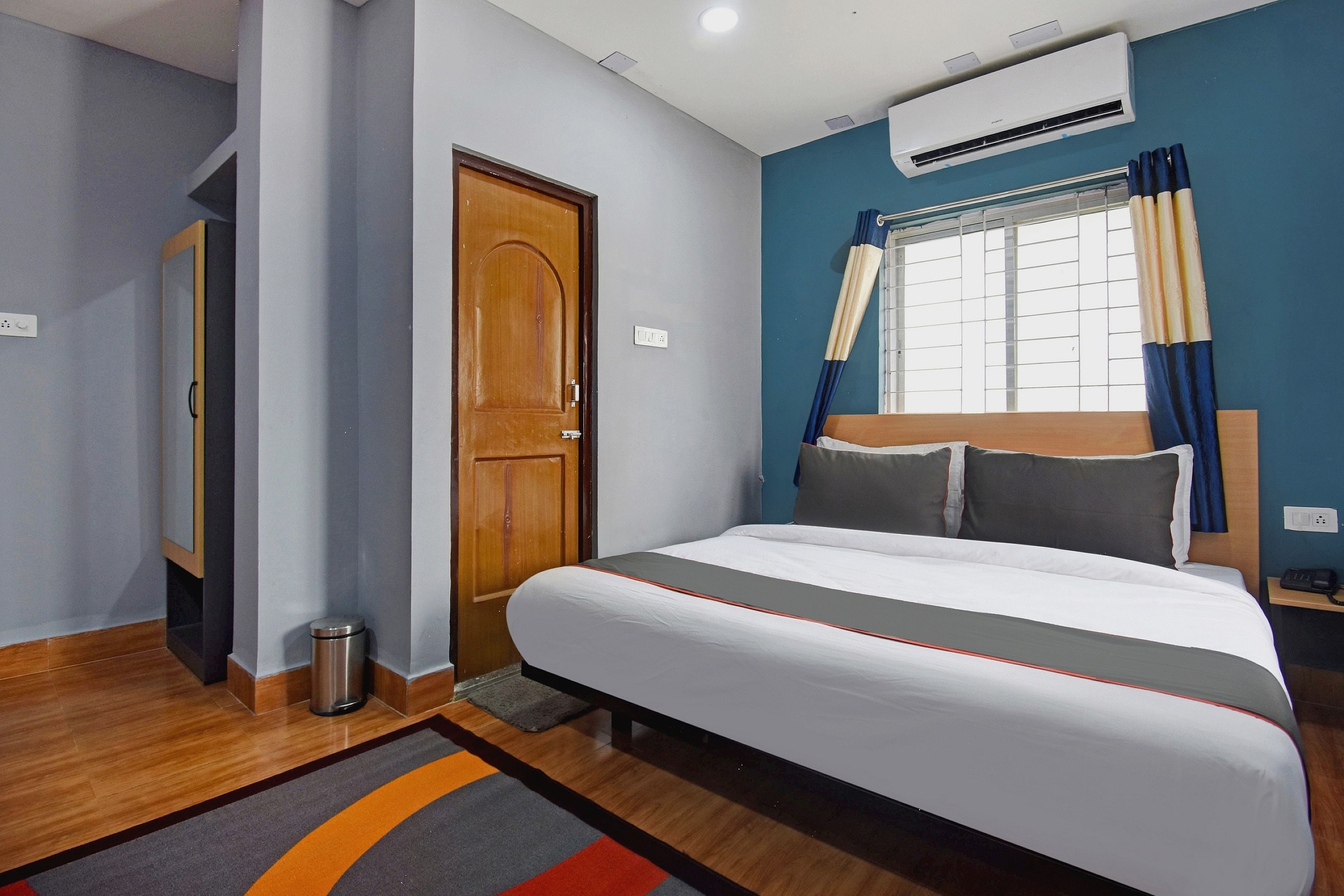 A hotel room with a bed, a wooden door, an air conditioning unit, a window with curtains, and a bedside table with a phone.
