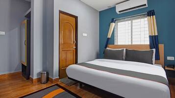 A hotel room with a bed, a wooden door, an air conditioning unit, a window with curtains, and a bedside table with a phone.