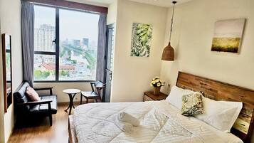 Une chambre d’hôtel dotée d’un grand lit, d’un coin salon avec un fauteuil et une petite table, d’une fenêtre donnant sur des bâtiments et d’œuvres d’art accrochées aux murs.