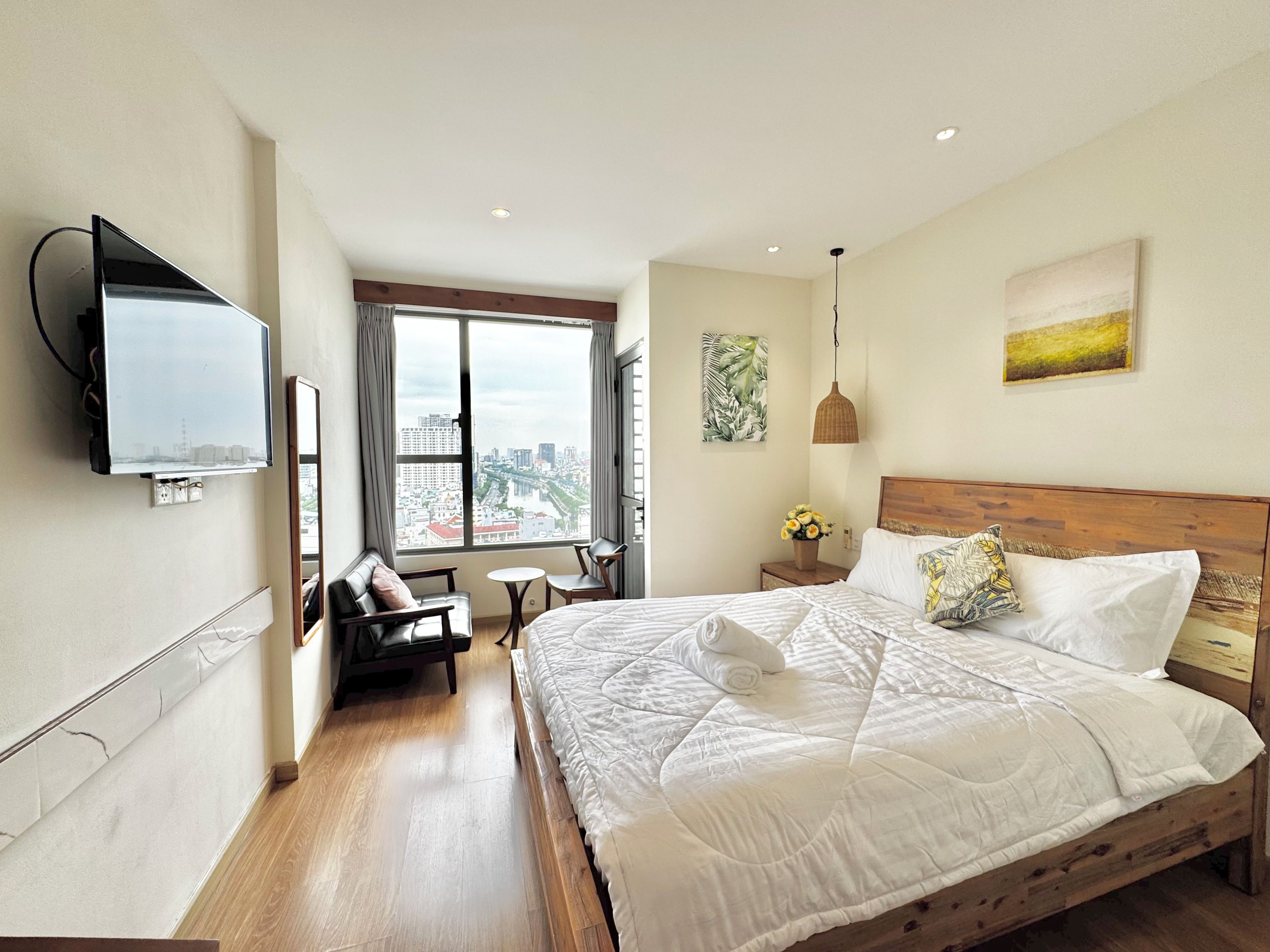 Habitación de hotel con una cama grande, un televisor montado en la pared, vistas a la ciudad a través de la ventana y una pequeña mesa con sillas.