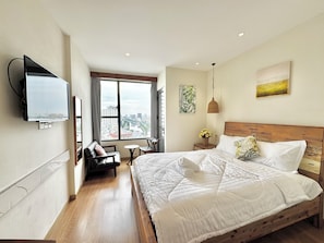 Habitación de hotel con una cama grande, un televisor montado en la pared, vistas a la ciudad a través de la ventana y una pequeña mesa con sillas.