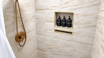 Zona de ducha con cabezal de ducha dorado y un rociador de mano. Hay un estante con tres botellas etiquetadas 'SHAMPOO,' 'CONDITIONER' y 'BODY SOAP'.