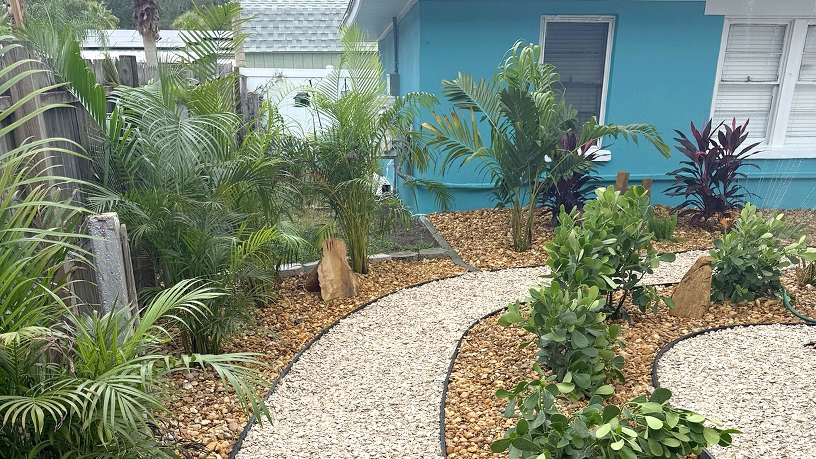 Un jardí amb un camí de grava, plantes diverses i un edifici blau al fons.