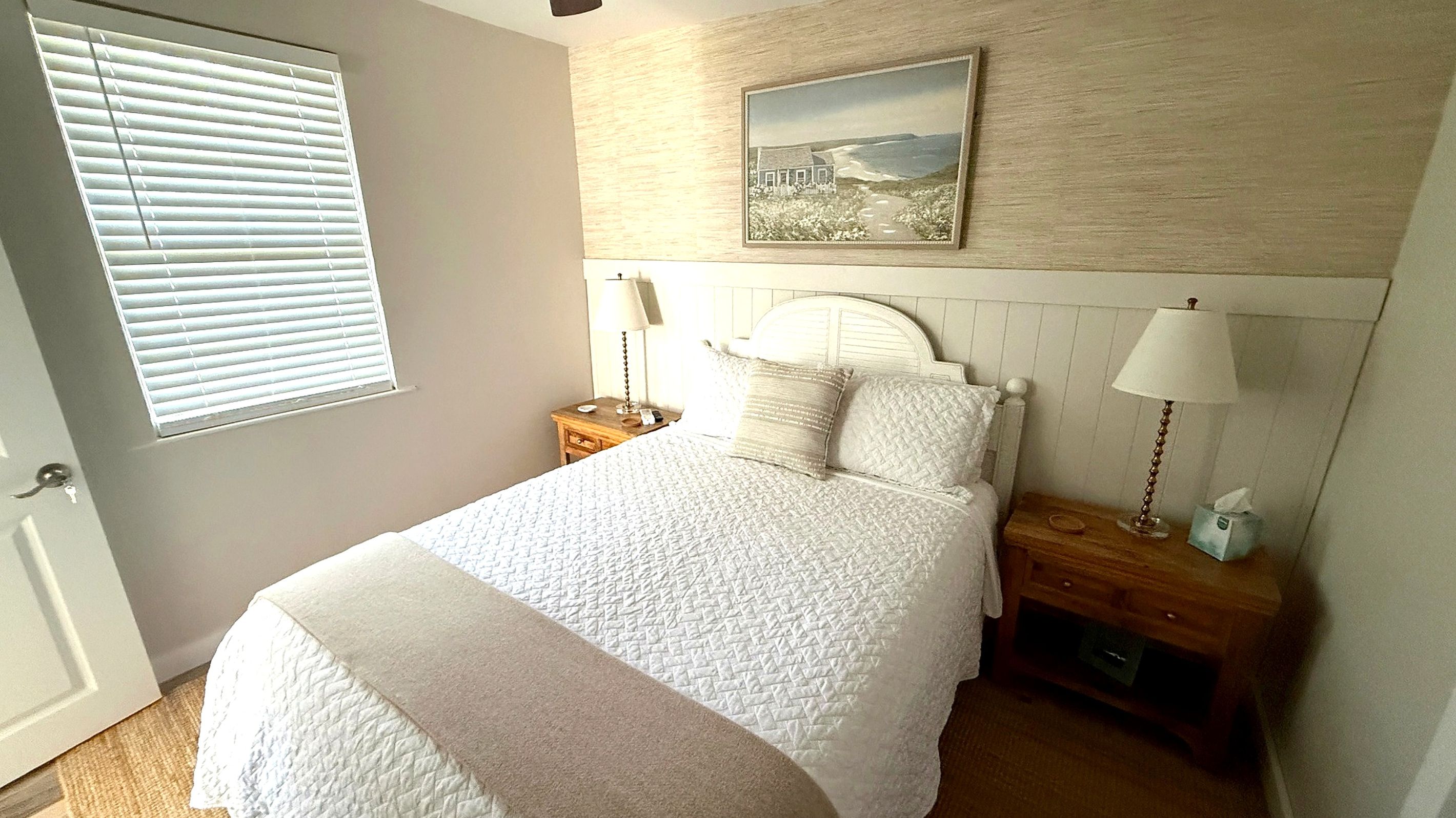 A bedroom with a bed, two bedside tables, a window with blinds, and a wall-mounted picture.