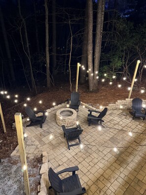 Un pati exterior amb un foc de pedra, seients Adirondack negres i llums penjants.