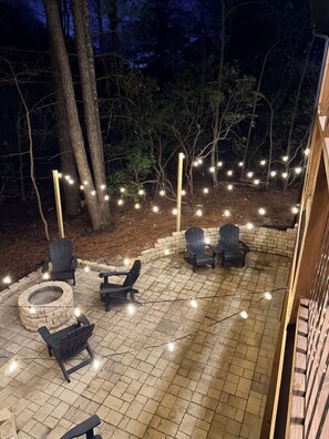 Ein Außenbereich mit einer Terrasse, Ziegelboden, Holzstühlen und einer Feuerstelle aus Stein.