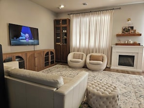 A living room with a sofa, two armchairs, a television, a cabinet, a fireplace, and a large window with curtains. - Beautiful & modern townhome! Master bedroom + two bedrooms. (Lakeville)